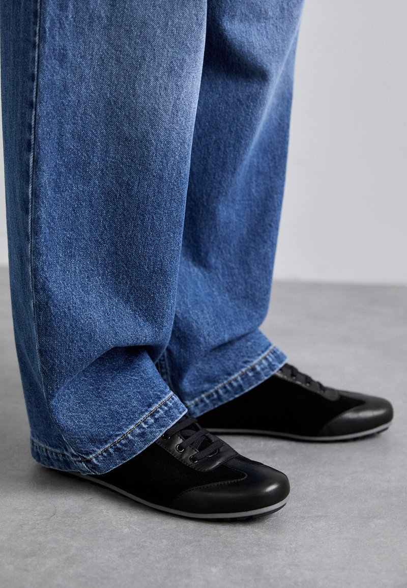 Black sneakers featuring a mix of smooth and suede textures, with laces and a contrasting rubber sole. Worn with loose blue denim pants.