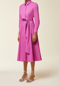 Abito camicia fucsia con colletto e maniche lunghe, caratterizzato da un dettaglio in vita annodato. Il modello indossa sandali bianchi con cinturini.