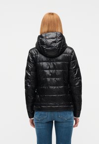 Γυναίκα με ίσια κόκκινα μαλλιά που φοράει μαύρο καπιτονέ μπουφάν με κουκούλα και μπλε τζι ν, στέκεται με την πλάτη προς τον θεατή.