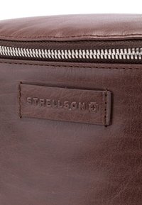 Rjava usnjena torba z reliefno površino, srebrnim zadrgo in vtisnjenim napisom "Strellson". Čiste linije in minimalističen dizajn.