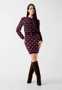 Robe-pull bordeaux avec un motif floral en losange, poignets côtelés et fermeture à boutons. Associée à des bottes en daim marron montant jusqu'aux genoux.