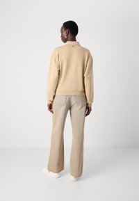 Felpa beige con una vestibilità rilassata, caratterizzata da un piccolo logo sulla parte posteriore, abbinata a jeans chiari a campana e sneakers bianche.