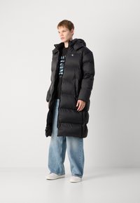 Lang sort pufferjakke med hætte, med et quiltet mønster, lynlåslommer og en afslappet pasform. Båret sammen med løse blå jeans og hvide sko.