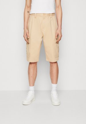 Beige cargoshorts met een ontspannen pasvorm, voorzien van zijzakken en een platte voorkant. Gecombineerd met witte sneakers en sokken.