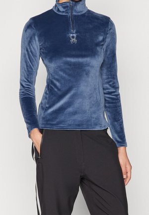 Kobieta ubrana w dopasowany długi rękaw z granatowego weluru z zamkiem z przodu i małym logo pająka, w połączeniu z czarnymi spodniami.