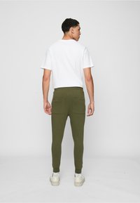 Grüne Baumwoll-Jogginghosen mit zwei Gesäßtaschen, taillierter Passform und elastischen Bündchen, kombiniert mit einem schlichten weißen T-Shirt. Modell von hinten betrachtet.