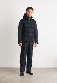 Black puffer jacket with a hood, featuring a zip front, rectangular quilted pattern, and smooth fabric. Worn with black trousers and shoes.