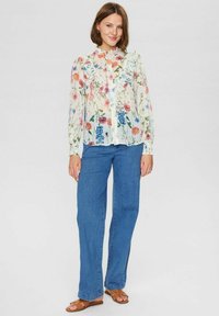 Blommig blus med volanger, med flerfärgade blommor på ett genomskinligt vitt material. Kombinerad med ljusblå utsvängda jeans och bruna sandaler.
