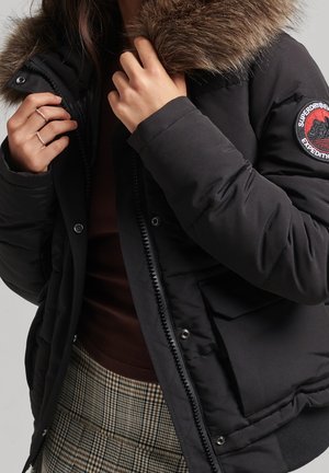 Woman wearing a black puffer jacket with fur collar, adjusting collar with hands, plaid skirt, and dark brown ribbed top visible.