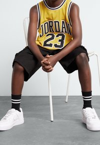 Żółta siatkowa koszulka do koszykówki z nadrukiem "JORDAN" i "23" na czarno, w połączeniu z czarnymi spodniami, skarpetkami w czarno-białe paski oraz białymi trampkami.