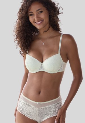 Smiling woman with curly hair wearing white lace bra and matching high-waist lace underwear, standing against light gray background.