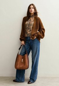 Brown knitted cardigan over a patterned blouse, paired with high-waisted blue jeans and a brown leather handbag. Dark pointed shoes complete the look.