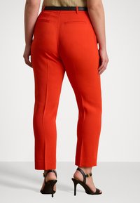 Pantaloni rossi su misura con vestibilità slim, dotati di tasche posteriori e una cintura nera. Tessuto liscio, taglio dritto, che termina poco sopra la caviglia.