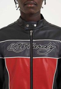 Sort læderjakke med røde og marineblå vandrette sektioner. Har hvid piping og broderet "Ed Hardy" logo på brystet, lynlås lukning og trykknapkrave.