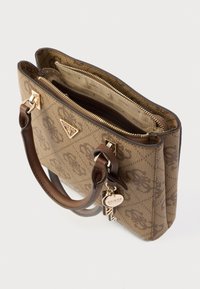 Ruda Guess rankinė su monogramos raštu, dviem rankenomis, auksiniu užtrauktuku ir firminiu pakabuku ant balto fono.