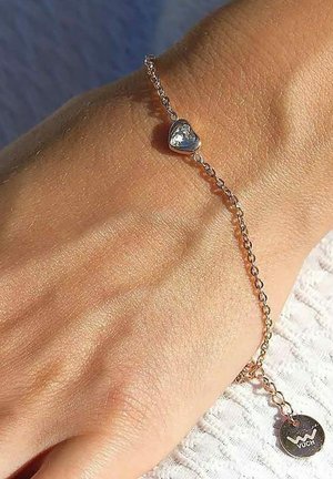 Silberne Armkette mit einem kleinen Herzanhänger, der einen klaren Edelstein enthält, befestigt an einem runden Anhänger mit der Gravur "VUCH".