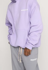 Felpa color lavanda con tasca frontale e logo "MULTIPLY" bianco, abbinata a pantaloni tuta grigi con logo abbinato e coulisse.