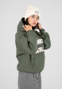 Joven ajustando la capucha de una sudadera verde con texto blanco, usando un gorro de punto crema y pantalones grises, contra un fondo claro y liso.