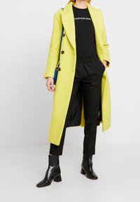 Kobieta w długim żółtym płaszczu, pod którym ma na sobie czarną koszulkę z dżinsów Calvin Klein, czarne spodnie i czarne botki za kostkę, trzymająca niebieską torebkę.