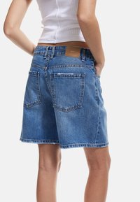 Denim shorts dengan pinggang mid-rise, warna biru, tepi yang robek, dua kantong belakang, dan patch kulit coklat pada pinggang.