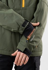 Olivgrön utomhusjacka med en gul accent, texturerat tyg, justeringar av kardborreband vid ärmarna och synligt varumärke på ärmen.