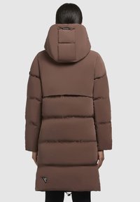 Pruun puffertagi, millel on kapuuts, eristuvad kudumisega paneelid, lahtine lõige ja reguleeritav nöör alumises servas. Diskreetne logo detail alumises servas.