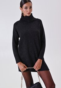 Zwarte gebreide jurk met hoge kraag, voorzien van fonkelende accenten. Het model houdt een tas met kettingband vast en draagt doorschijnende zwarte panty’s.