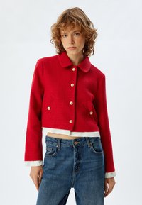 Rote, strukturierte, verkürzte Jacke mit rundem Kragen, goldenen Knöpfen und zwei vorderen Taschen; getragen über einem weißen, geschichteten Shirt und blauen Jeans.