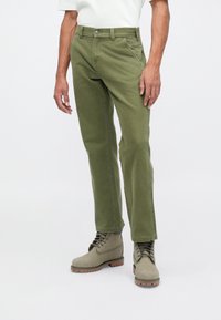 Homem com calças straight-leg verde-oliva e botas de atacadores combinando, de pé contra um fundo claro e liso.