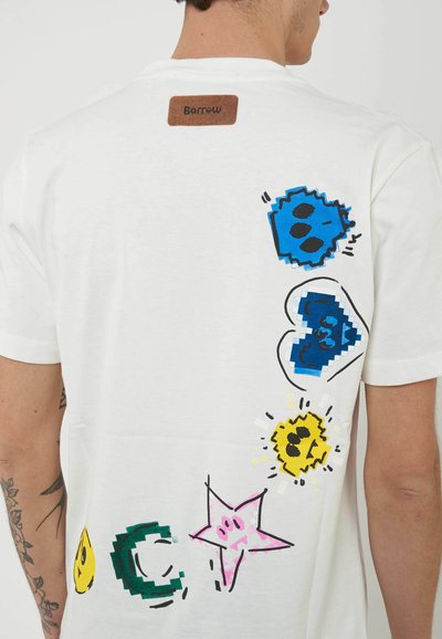 T-shirt bianca con forme pixelate colorate tra cui teschi blu, un cuore blu, sole giallo, stella rosa, mezzaluna verde e goccia gialla lungo la schiena.