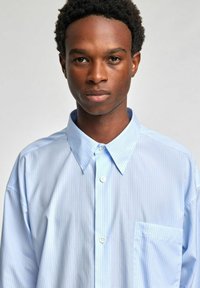 Lys blåstripet skjorte med vid krage, knappelukking og en lomme med en subtil logo. Glatt stoff med en avslappet passform.