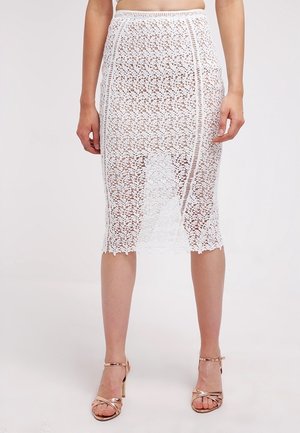 Kobieta ubrana w białą, koronkową spódnicę do kolan z kwiatowymi wzorami oraz beżowe sandały na wysokim obcasie, stojąca na jednolitym tle.