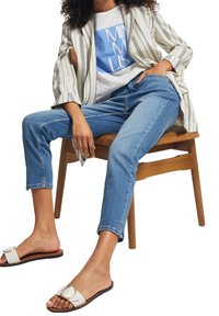 Blazer a rayas, camiseta blanca con gráfico azul, jeans de mezclilla azul claro y sandalias blancas de tiras con un acento circular, sentado en una silla de madera.