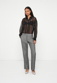 Mulher vestindo blusa preta transparente com botões, sutiã preto, calças xadrez cinza e saltos pontiagudos bege, em pé contra fundo branco.