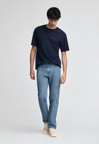 Marinblå t-shirt med kort ärm och texten "Polo Club", ljusblå raka jeans, och beige sneakers på en modell som står framför en neutral bakgrund.
