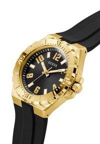 Gullfarget klokke med svart urskive, store fete timemarkører, teksturert bezel og matt svart gummirem. Har en kronen på siden.