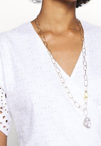 Collana a catena d'oro con un pendente in oro texture e un accento di perla dalla forma irregolare, indossata sopra una blusa bianca in pizzo.