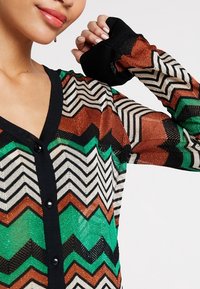 Närbild av person som bär en flerfärgad zigzag-mönstrad kofta med svarta knappar och svarta detaljer, armen höjd nära ansiktet.