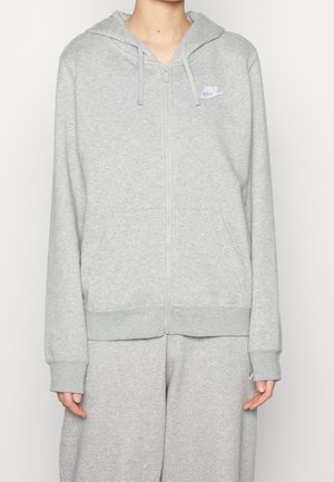 Personne portant un sweat à capuche zippé Nike gris clair et un pantalon de survêtement assorti, debout devant un fond uni clair.