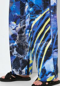 Blauwe en zwarte gestreepte wijde broek met abstracte patronen en gedurfde gele strepen; de stof lijkt glad te zijn. Gemaakt met zwarte gevlochten sandalen.