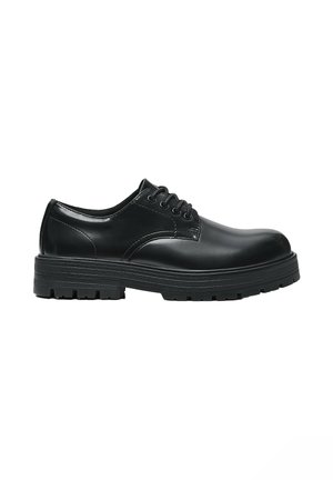 Zwarte leren veterschoen met dikke geribbelde zool en afgeronde neus, zijaanzicht op witte achtergrond.