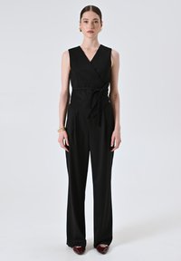 Completo senza maniche nero con design avvolgente, vita con cintura e pantaloni a gamba larga. Tessuto morbido senza fantasie. Paio di scarpe rosse.