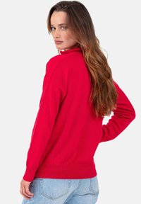 Pull en tricot rouge avec un col montant, des manches longues et un ourlet côtelé. Le tissu semble doux et texturé, associé à un jean bleu clair.