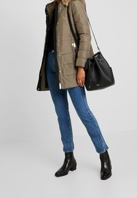 Olijfgroene gewatteerde jas met een met bont afgewerkte capuchon, gecombineerd met rechte blauwe jeans en zwarte enkellaarsjes. Zwarte bucket bag met zilverkleurige accenten.