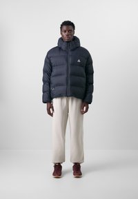 Svart dunjacka med hög krage, dragkedja framtill och triangulärt märke. Bärs med cremefärgade sweatpants och mörka sneakers.