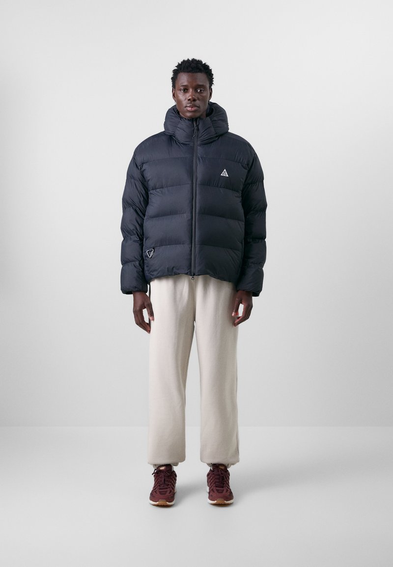 Svart dunjacka med hög krage, dragkedja framtill och triangulärt märke. Bärs med cremefärgade sweatpants och mörka sneakers.