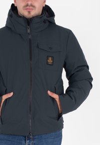 Ciemnoniebieska zimowa kurtka z kapturem, na zamek błyskawiczny, z przednią kieszenią i naszywką z logo; cechuje się gładką fakturą i elastycznymi mankietami.