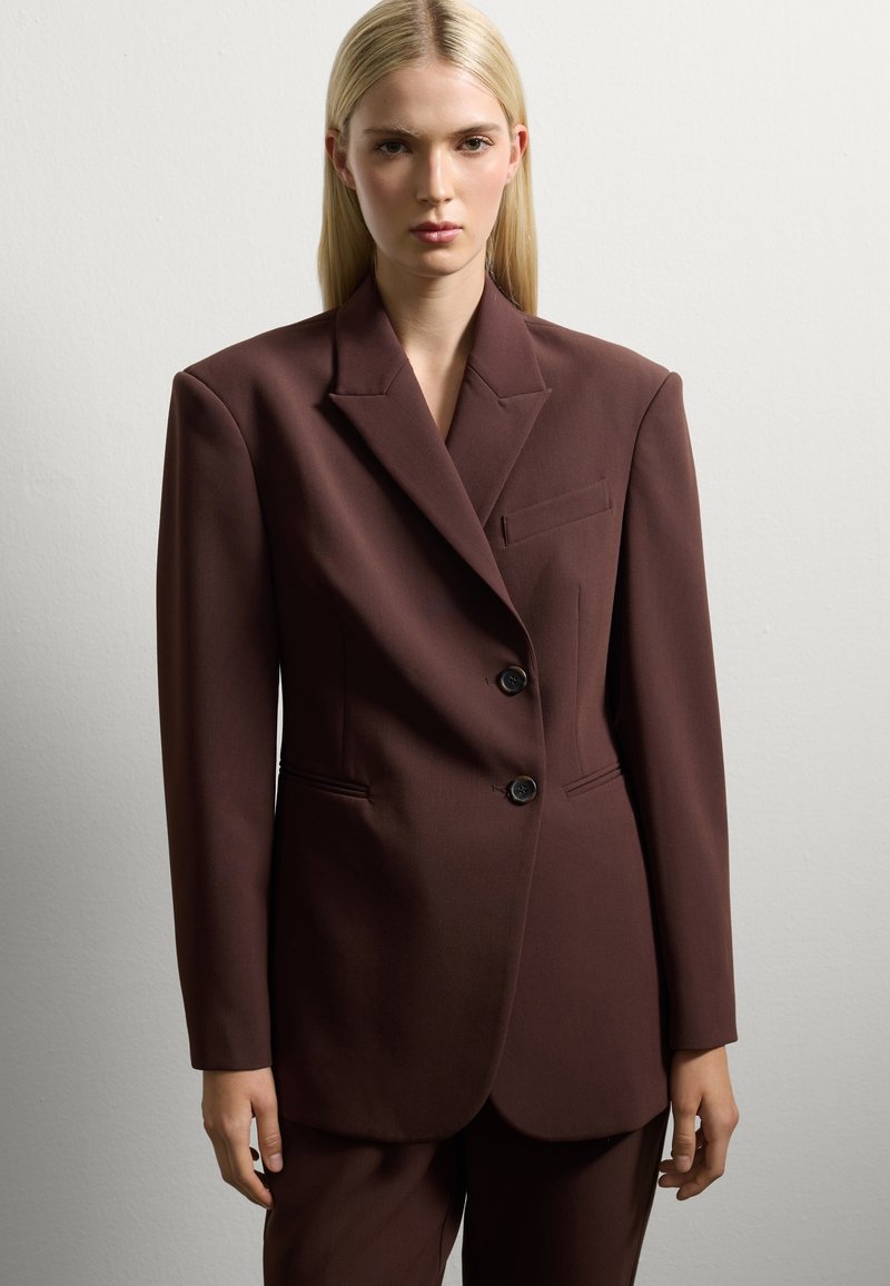 Blazer marrón a medida con solapas afiladas, dos botones y bolsillos laterales. Tejido suave con hombros estructurados.
