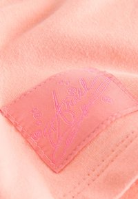 Primer plano de tela rosa que muestra un parche rectangular cosido con texto en cursiva y puntos decorativos sobre un material suave y texturizado.