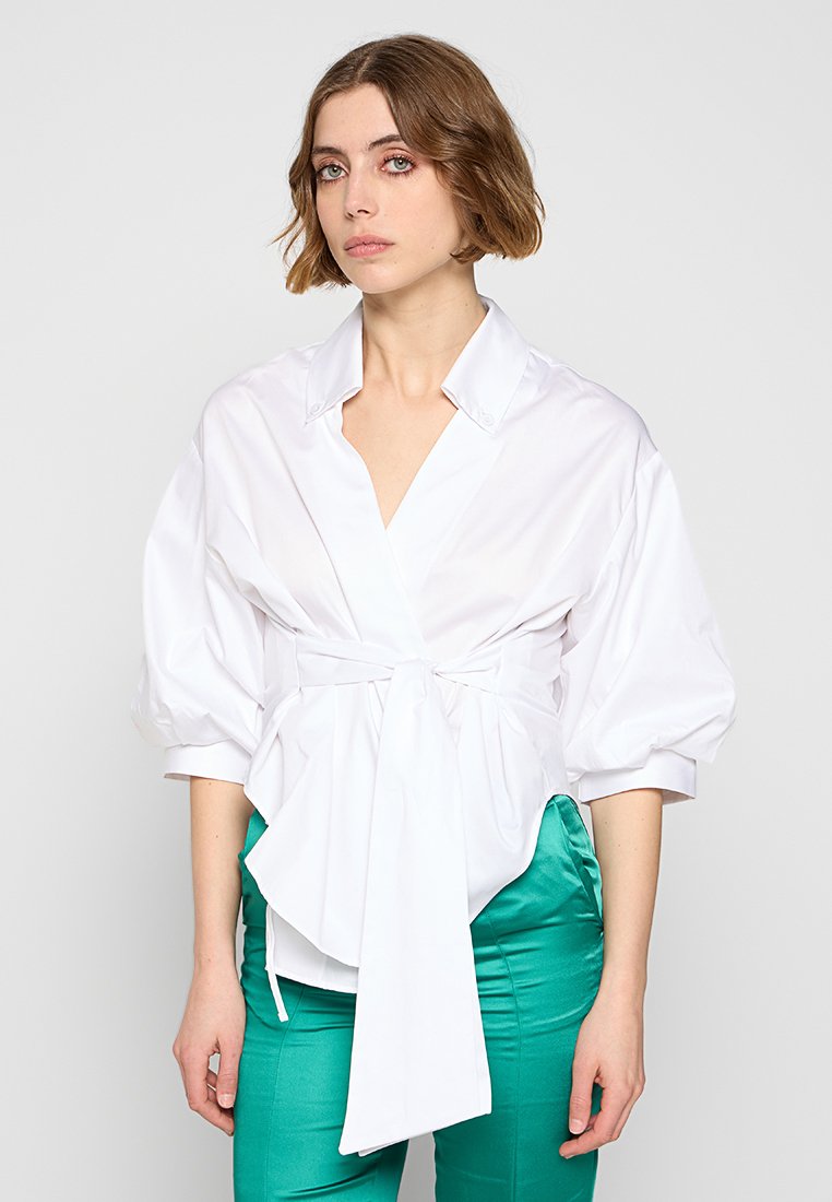 Weißes, kragenloses Hemd mit Puffärmeln, Wickeldesign und vorderer Schleifendetail. Glatte Textur. Kombiniert mit grünen Satin-Hosen.
